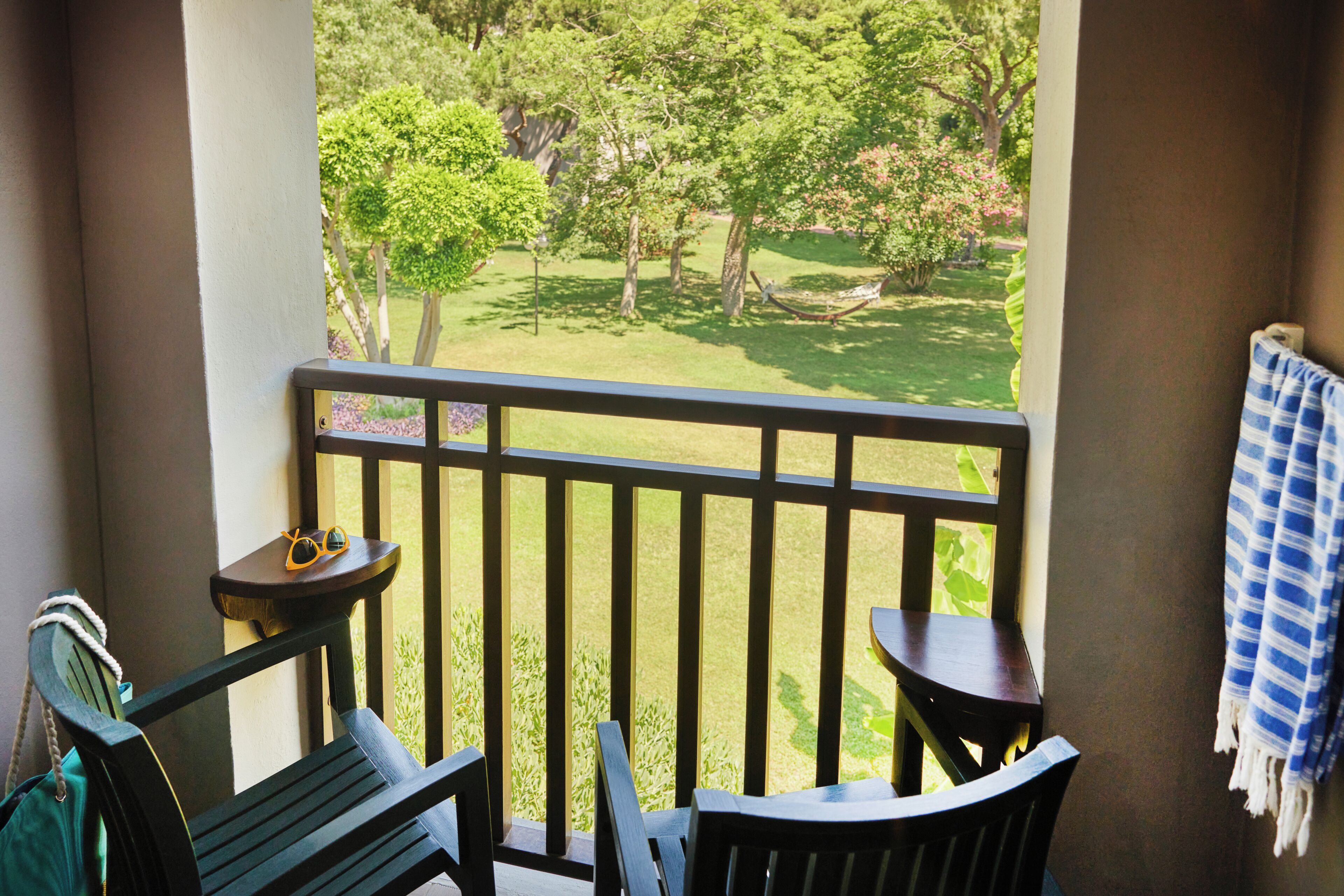 A balcony with a view of a grassy area, a hammock, a table, and a chair.