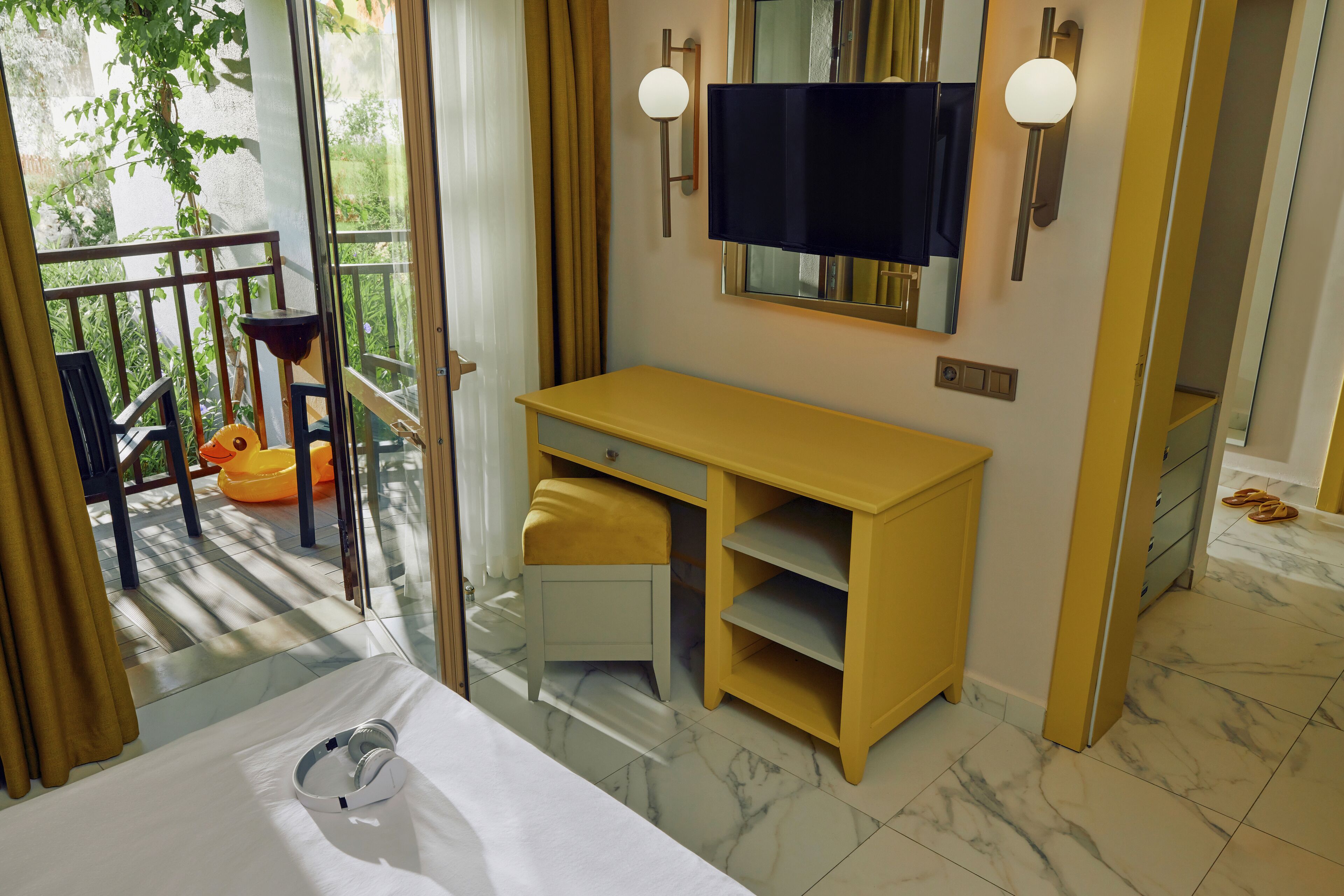 Une chambre d’hôtel comprenant un lit, un bureau, une chaise, un petit tabouret, une télévision et une fenêtre avec des rideaux.