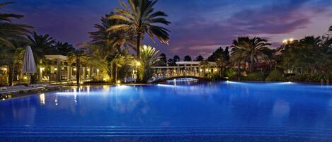 A large, illuminated swimming pool at dusk with palm trees and a bridge.