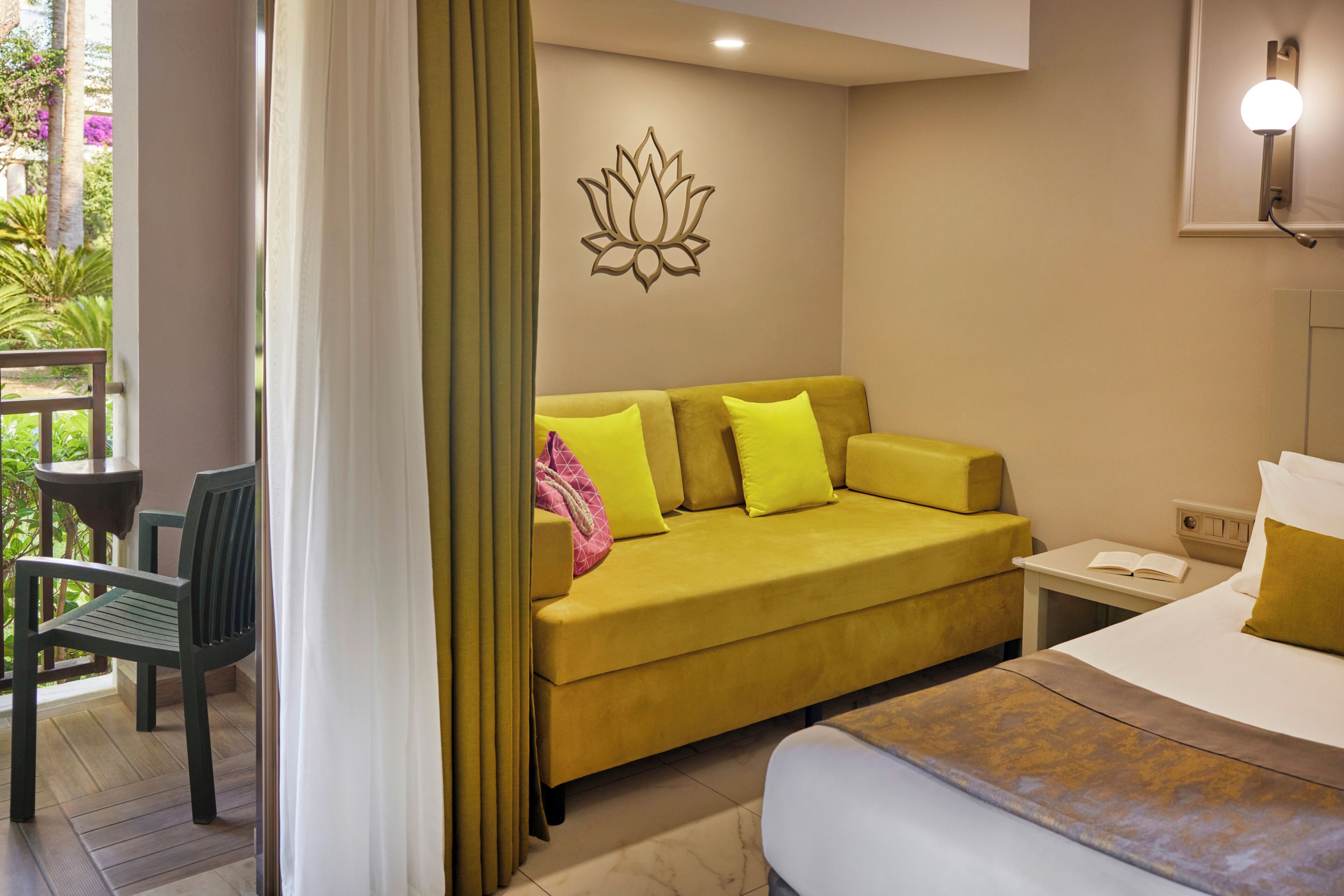 A hotel room with a yellow sofa, a bed, a nightstand, and a view of greenery outside.