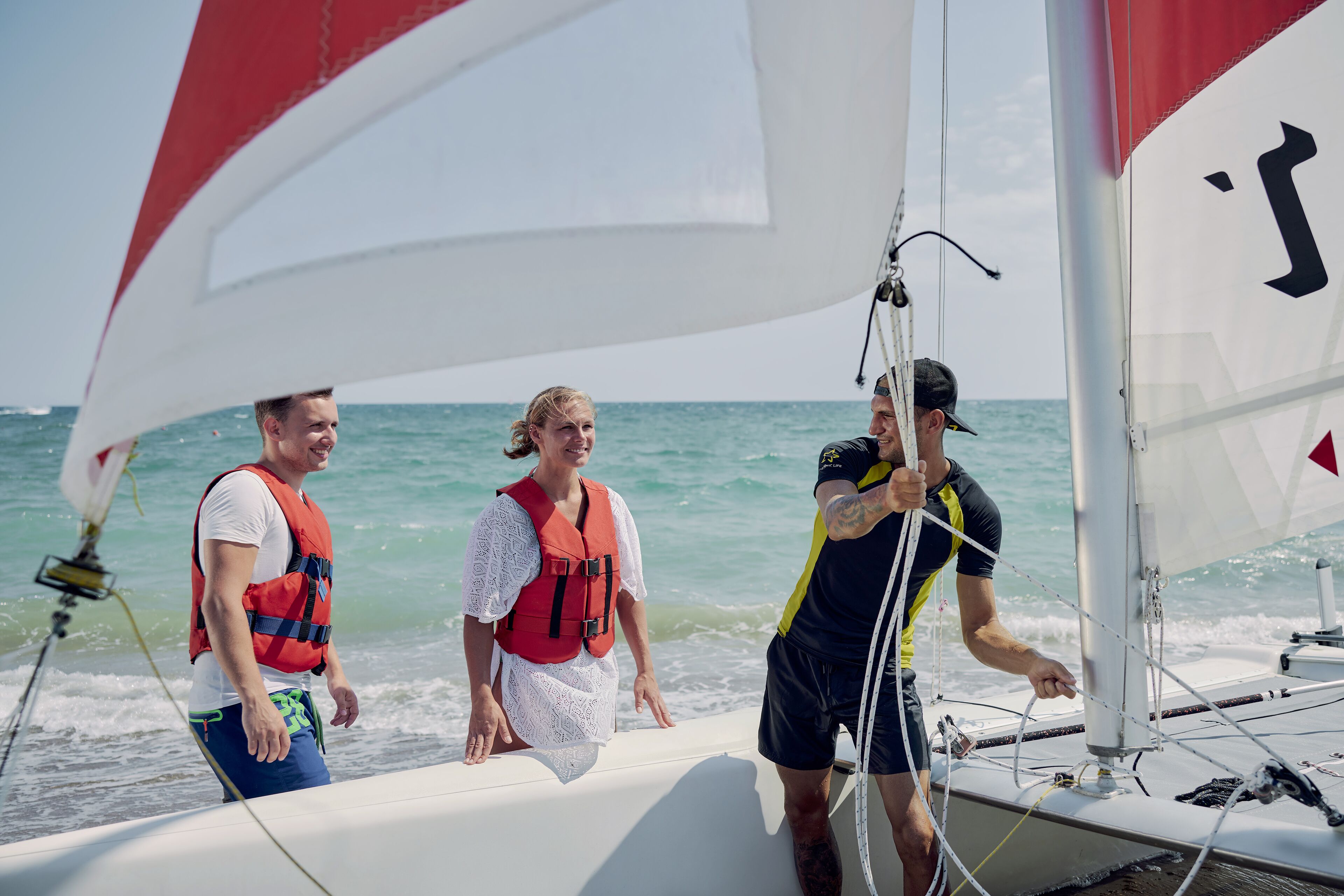 Three people on a sailboat with a white sail and a red stripe, wearing life jackets.