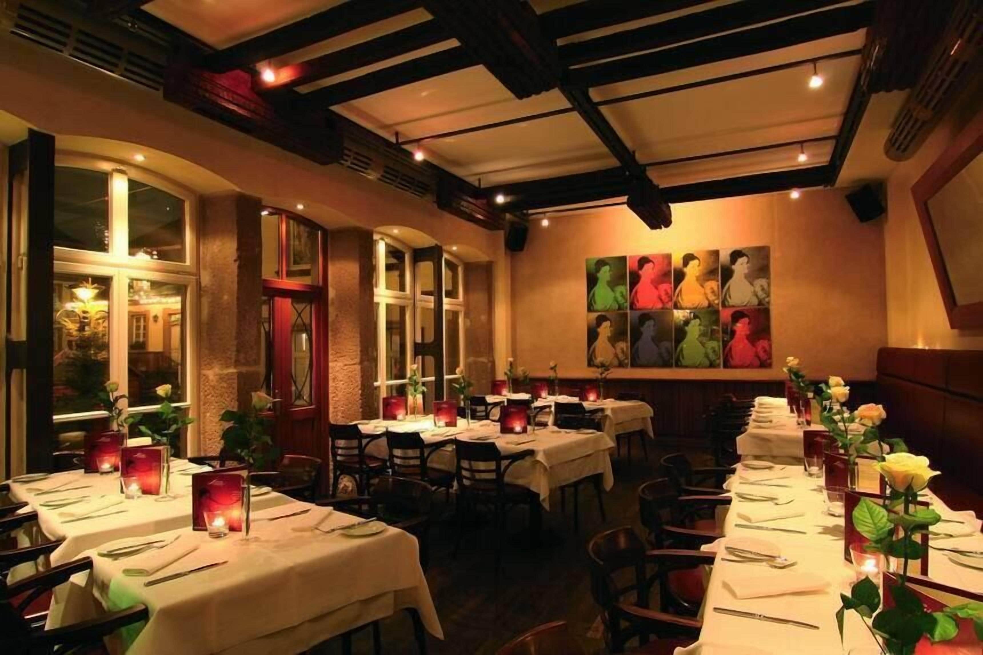 Un restaurante con largas mesas dispuestas para comer, con centros de mesa iluminados por velas y una fila de cuadros en la pared.