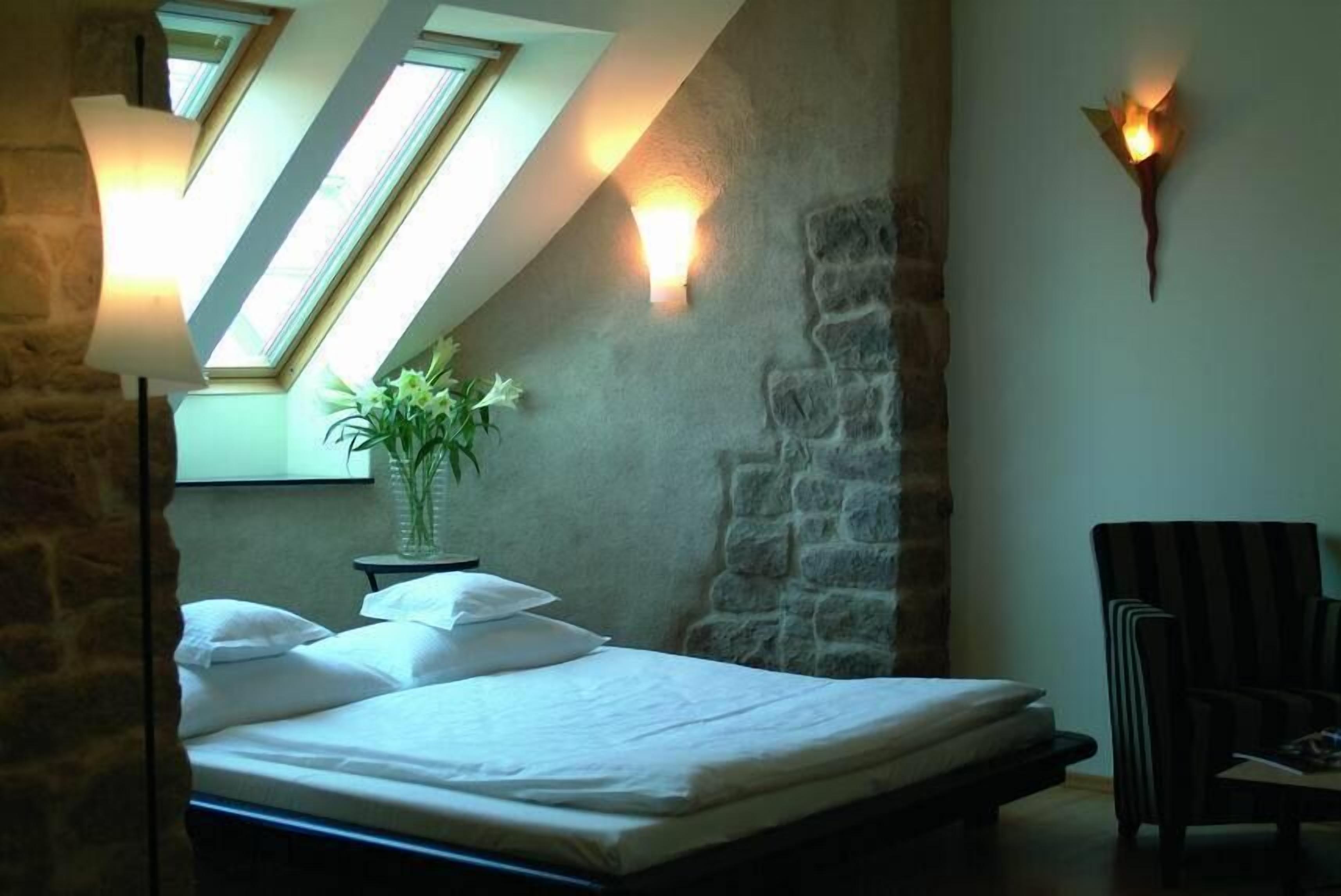 Quarto com teto inclinado, claraboias, cama, cadeira e luminária fixada na parede.