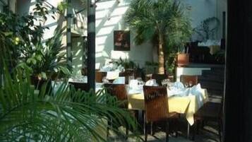 An outdoor dining area with tables and chairs, surrounded by greenery and a glass enclosure.