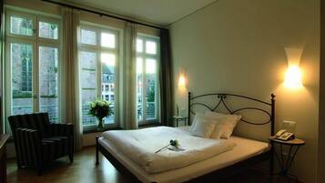 A hotel room with a large bed, a chair, a bedside table with a lamp, and a window with curtains.