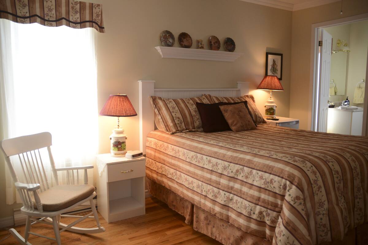 Un dormitorio con una cama, una mecedora, una mesita de noche y un estante con objetos decorativos.