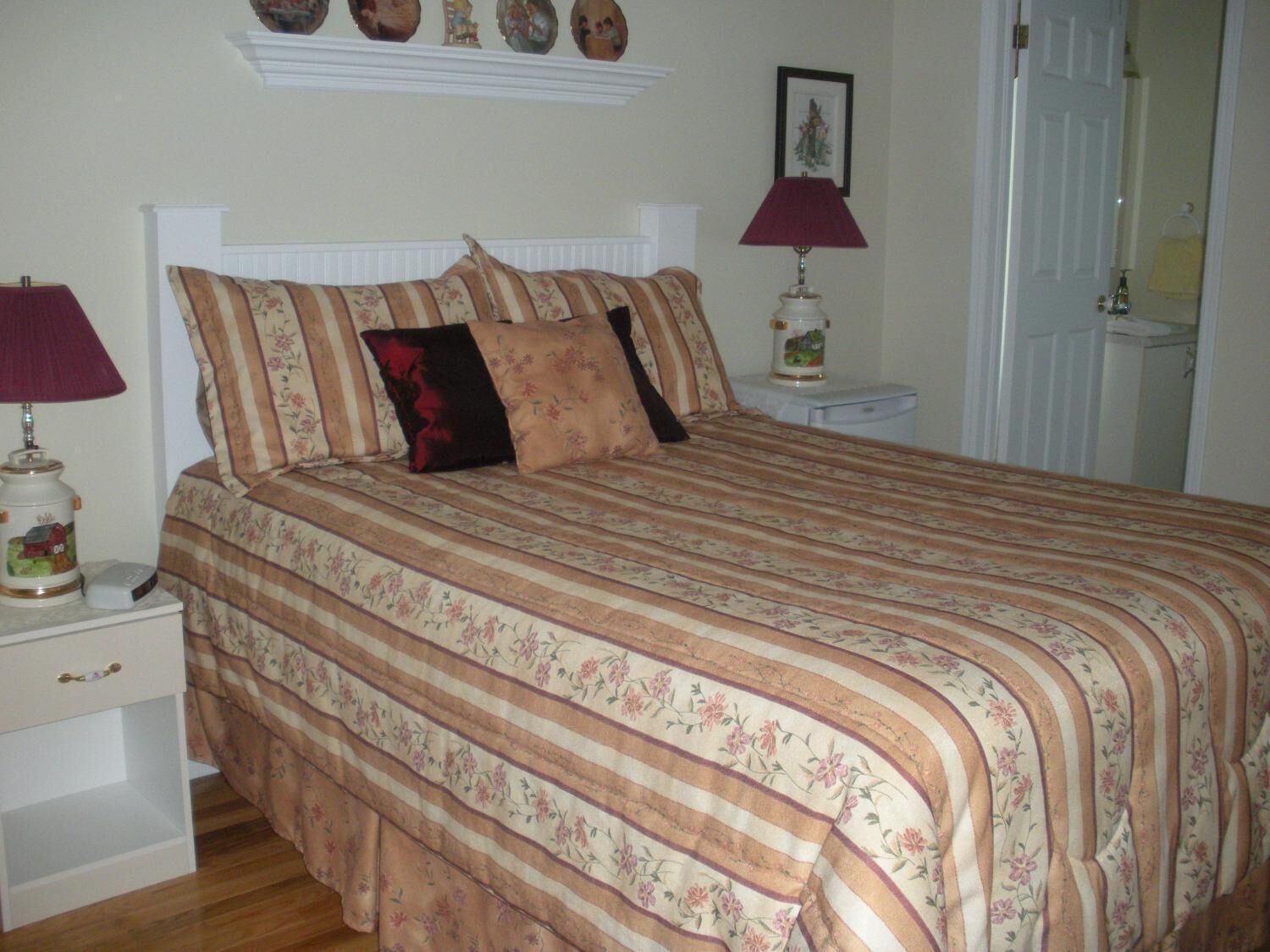 A bedroom with a bed, two bedside tables, a nightstand, a lamp, and a wall-mounted shelf with decorative items.