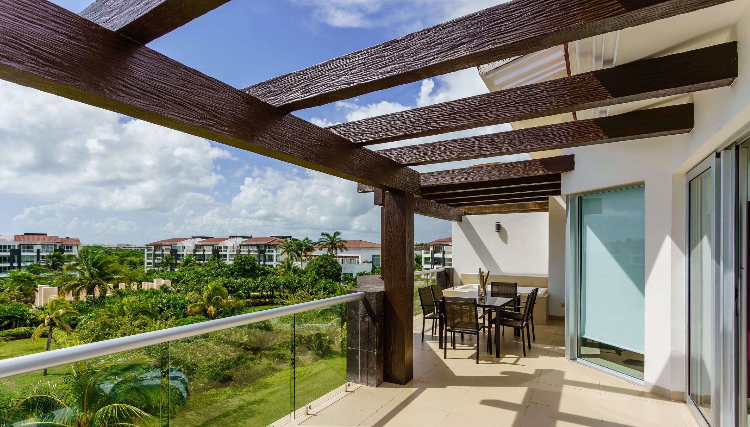 A balcony with a pergola, a dining area with chairs and a table, and a view of a residential complex with palm trees.