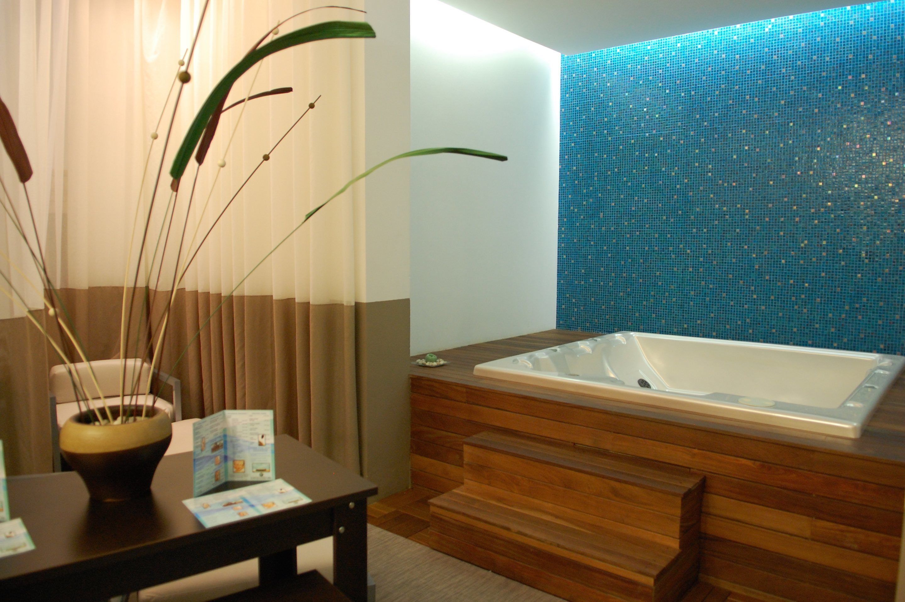 A modern bathroom with a wooden bathtub, a blue mosaic wall, and a table with a vase of plants.