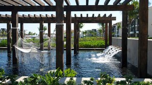 Eine Hängematte unter einer Holzpergola über einem Wasserspiel mit einem Wasserfall, umgeben von viel Grün und einem Pool.