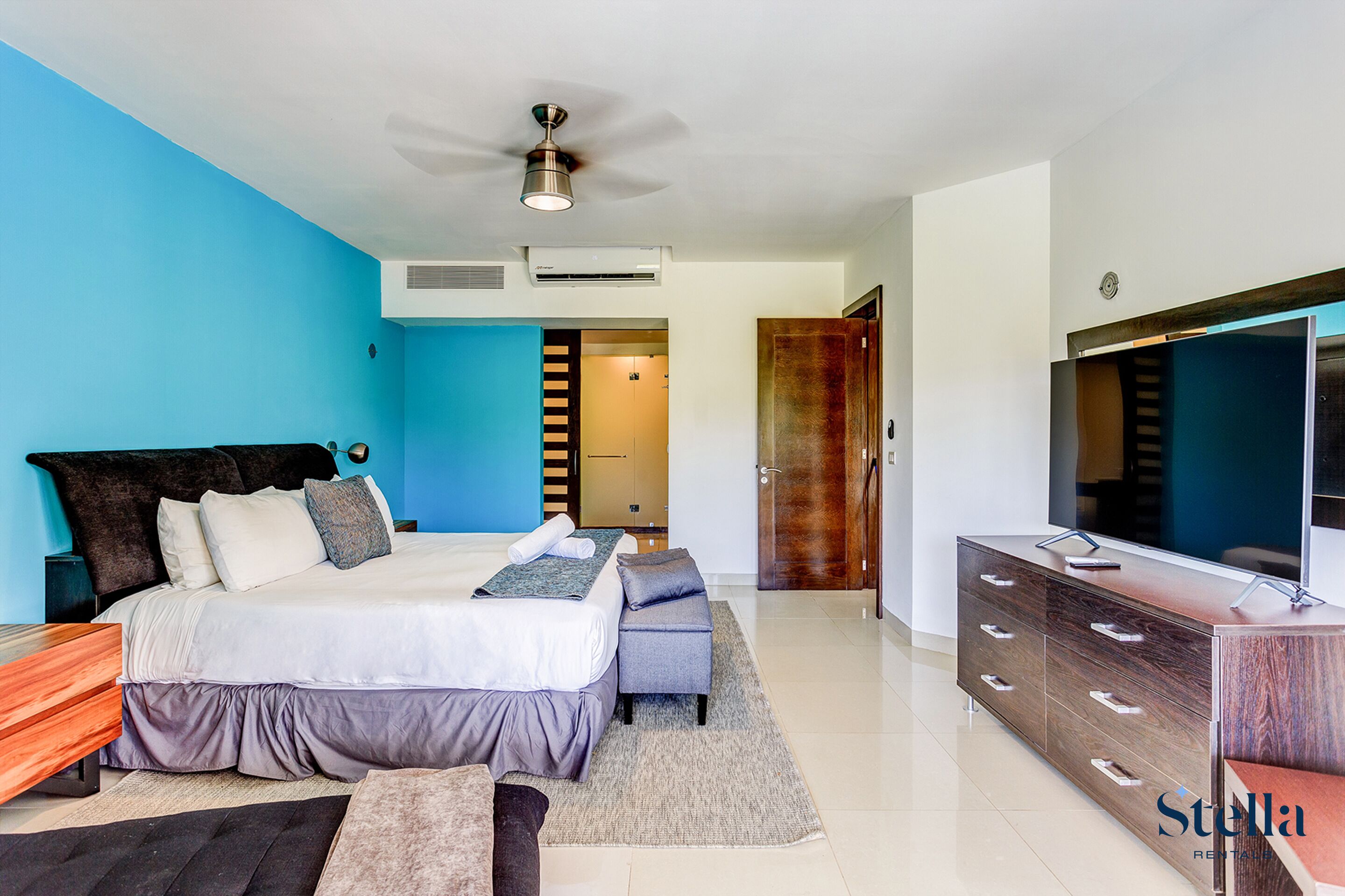 Une chambre à coucher moderne avec un grand lit, une télévision, une commode en bois et un ventilateur de plafond.