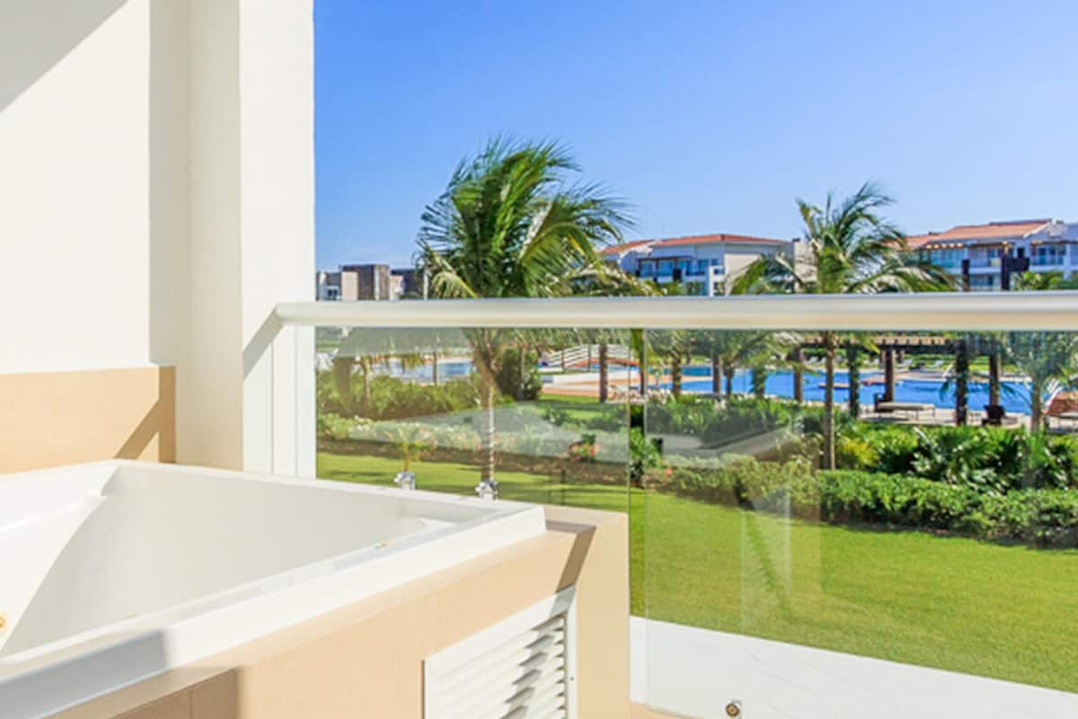 Ein modernes Badezimmer mit einer großen Badewanne, Blick auf ein Schwimmbad und Palmen durch ein Fenster.