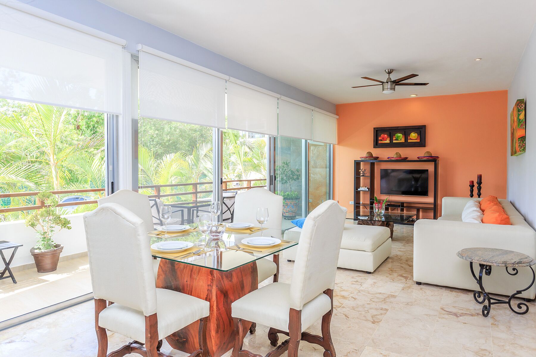 A spacious living area with a glass dining table, white furniture, a TV, and a balcony with a view of greenery.