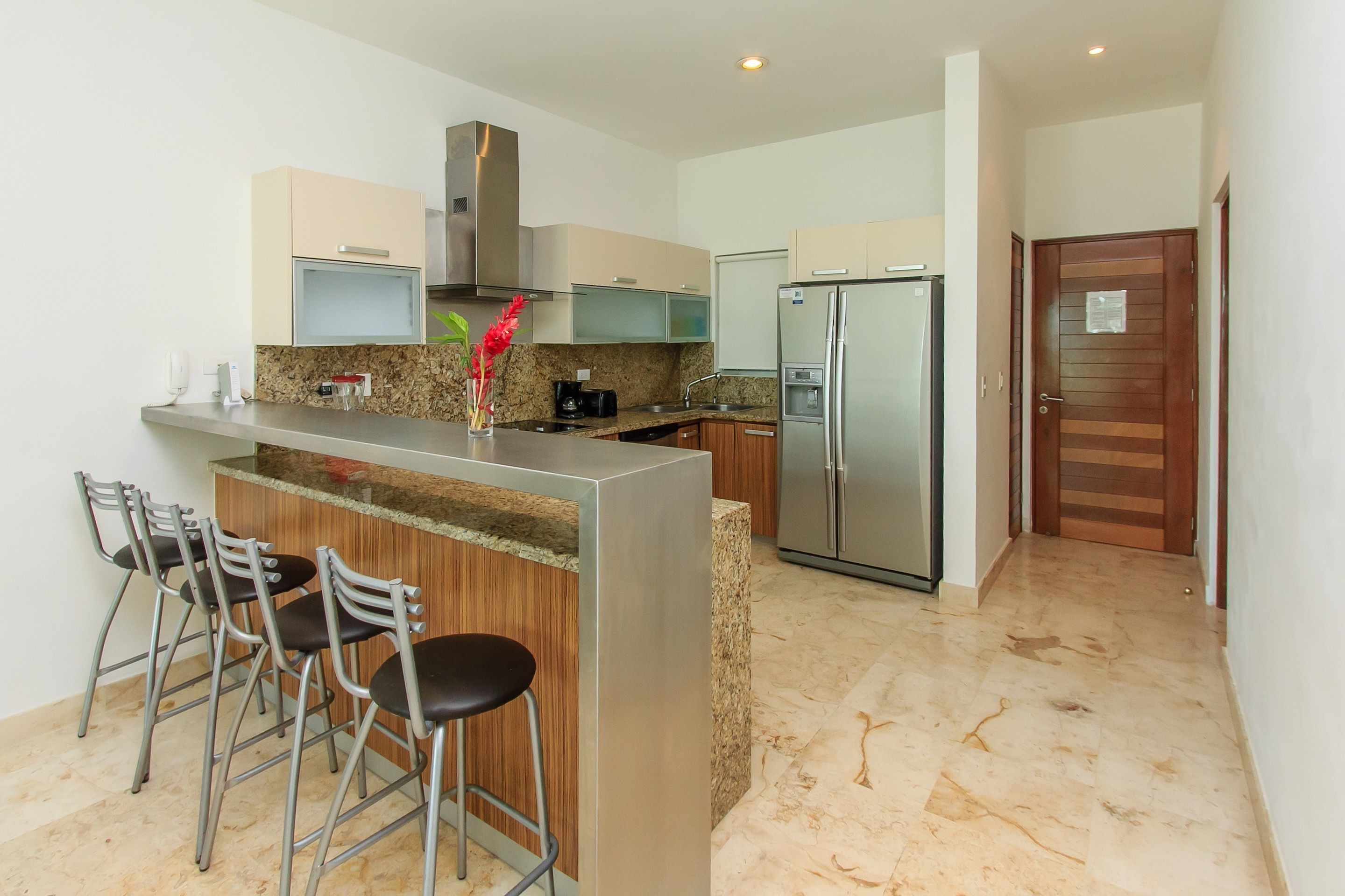 A modern kitchen with a central island, bar stools, stainless steel appliances, and a wooden door.