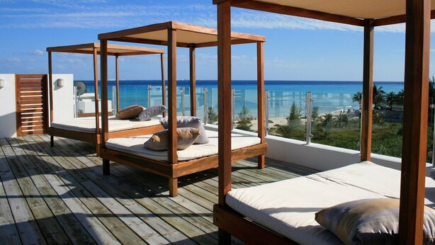 Un pontile in legno con due letti di legno, biancheria e cuscini, con vista su un mare limpido e una spiaggia.