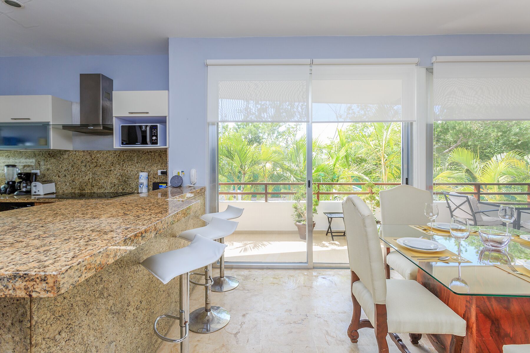 A modern kitchen with granite countertops, white bar stools, and a dining area with a glass table and white chairs.