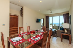 A dining area with a glass table set for six, wooden chairs, and a view of the ocean through large windows.