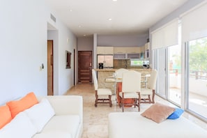 A modern living room with a sofa, a dining area with chairs and a table, a kitchen with cabinets and appliances, and a sliding glass door leading to a balcony.