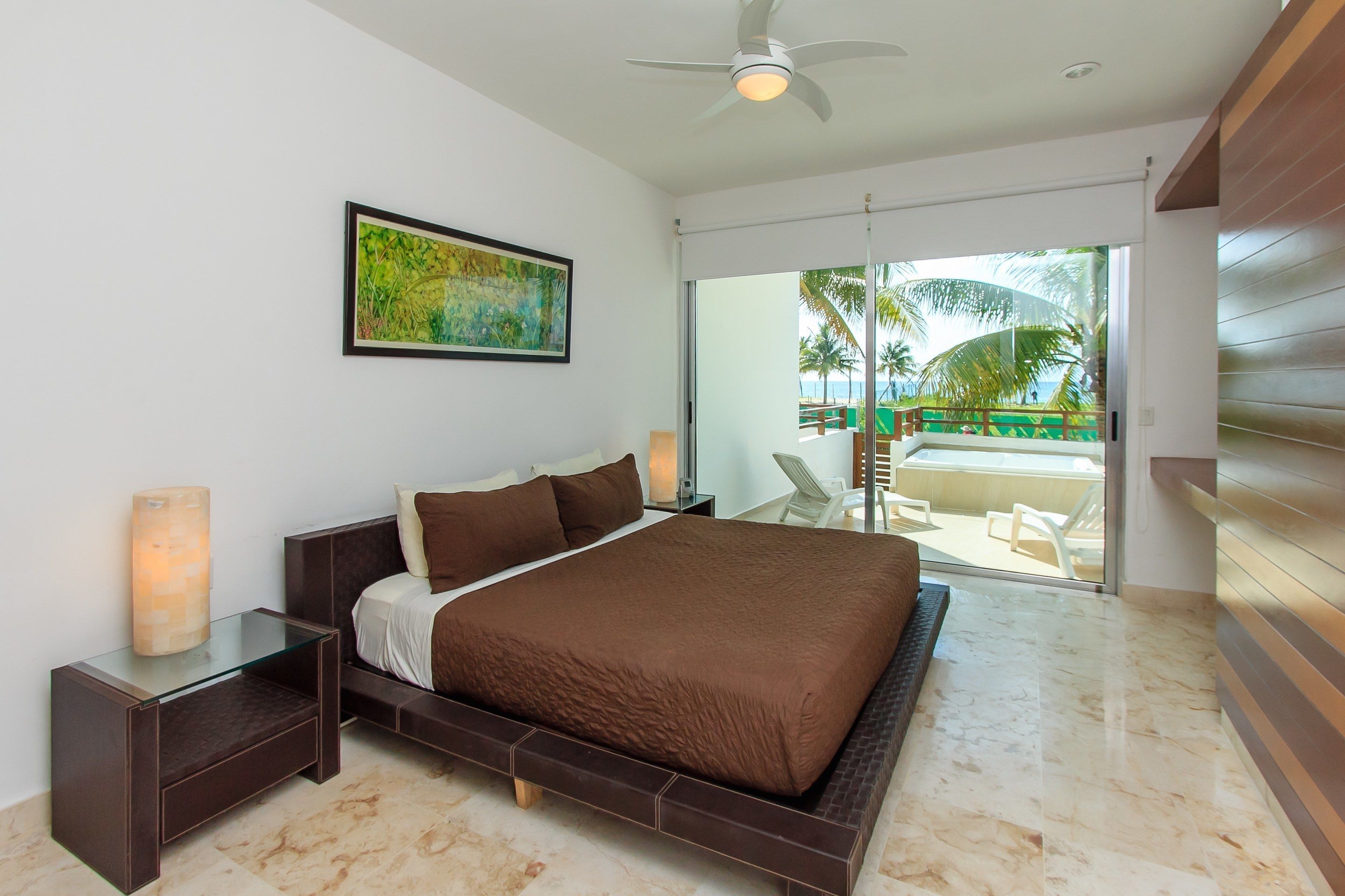 A modern bedroom with a large bed, a glass side table, a ceiling fan, and a view of a pool and palm trees through a sliding glass door.
