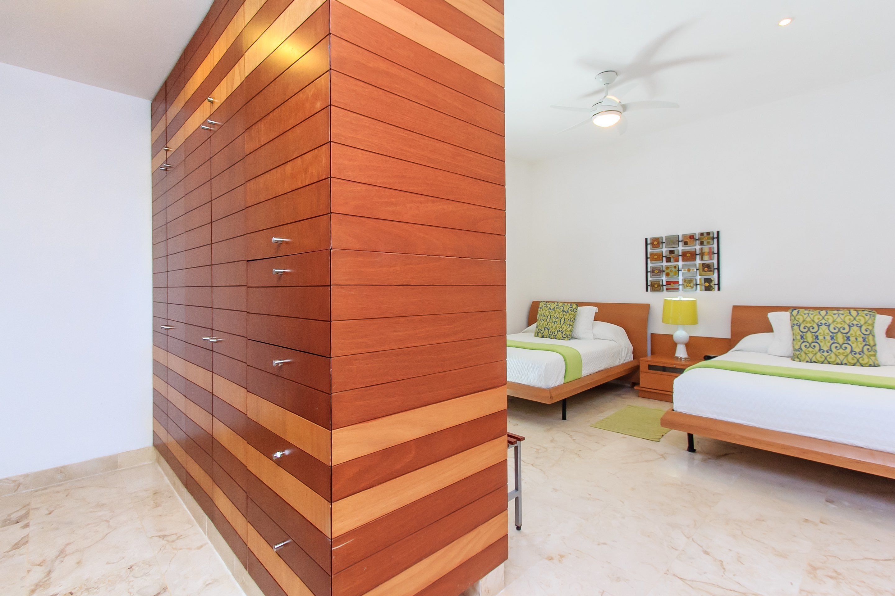 A modern bedroom with a wooden wardrobe, two single beds, a ceiling fan, and a wall-mounted artwork.