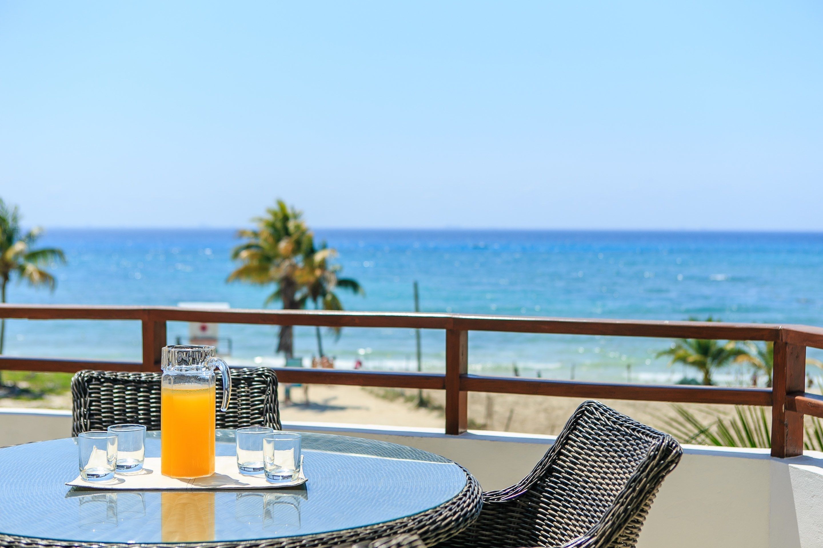 A table with a pitcher of orange juice and glasses, overlooking a beach with palm trees and the ocean.