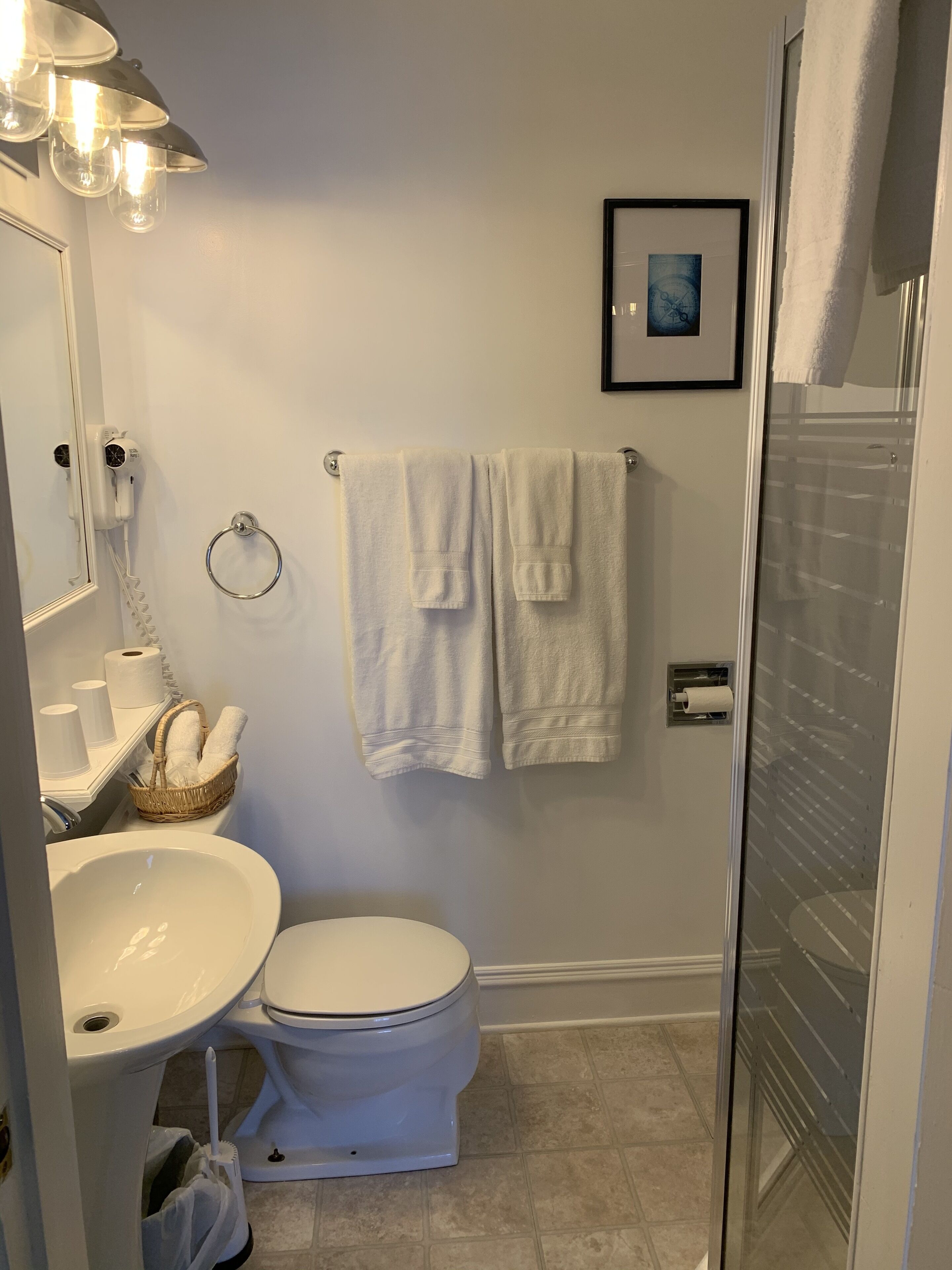 Un baño con inodoro, lavamanos y zona de ducha. Hay un perchero con dos toallas, un cuadro en la pared y un cesto con toallas sobre el lavamanos.