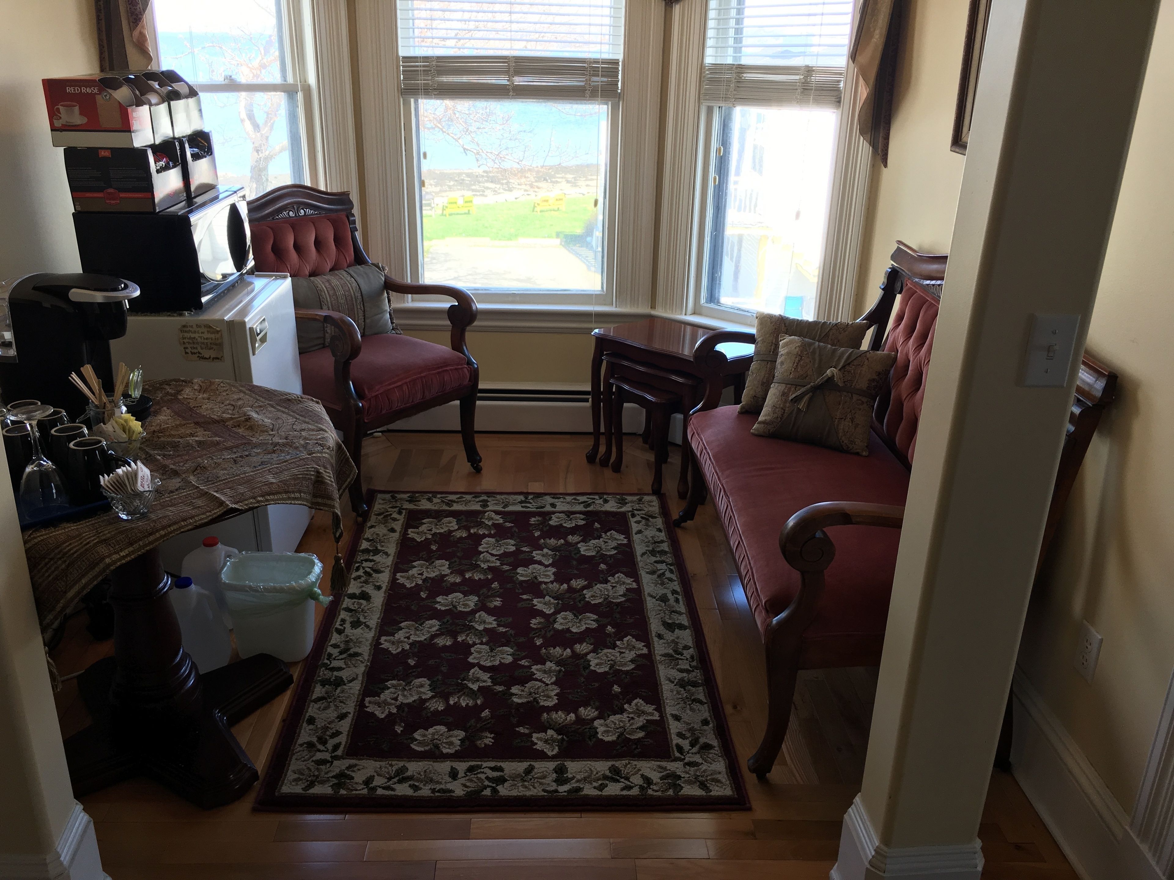 A cozy living room with a red sofa, a wooden coffee table, and a floral rug. There is a window with blinds, a small table with a vase, and a red chair.
