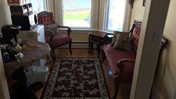 A cozy living room with a red sofa, a wooden coffee table, and a floral rug. There is a window with blinds, a small table with a vase, and a red chair.