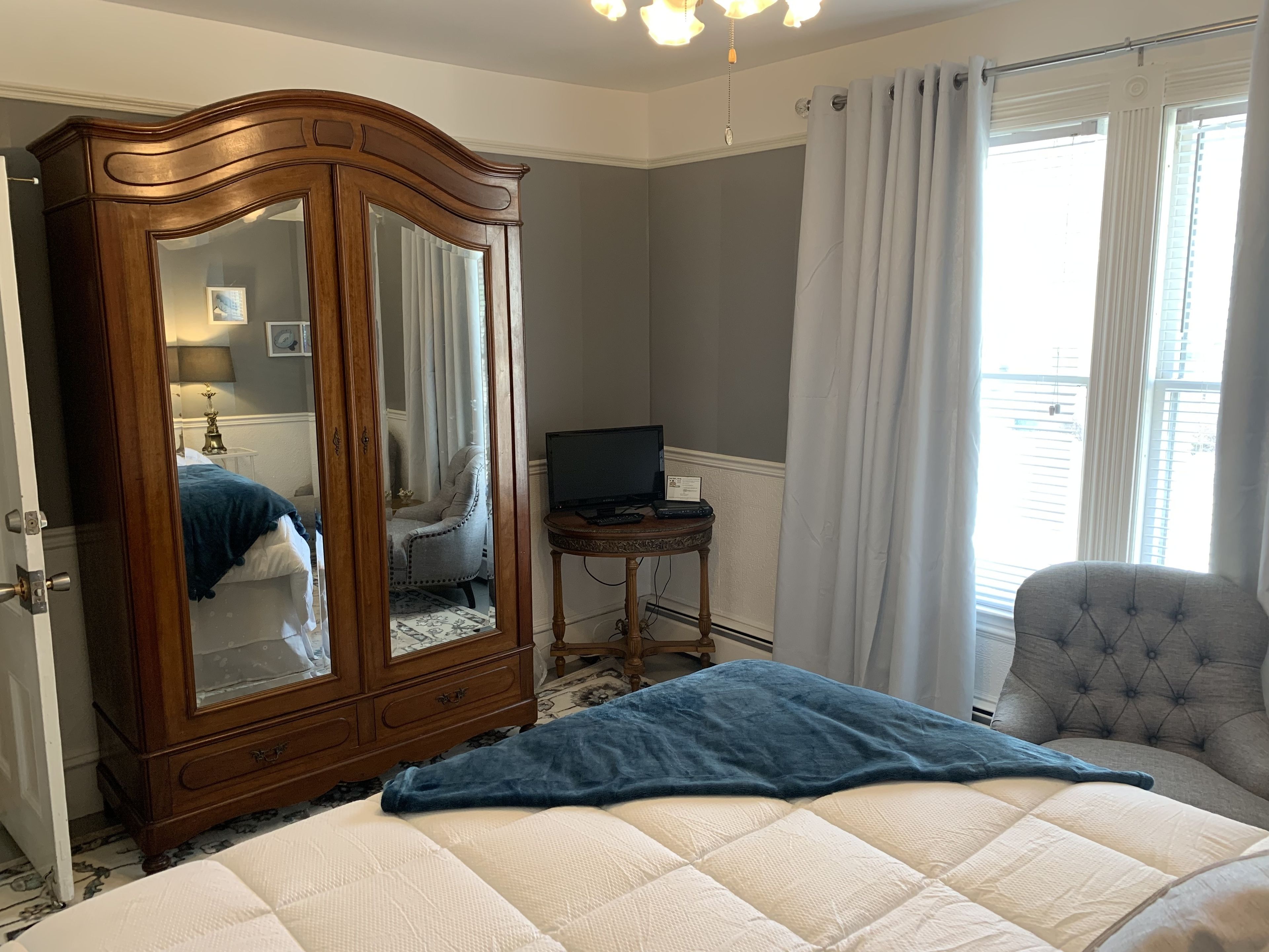 A bedroom with a large bed, a wooden wardrobe, a small table with a television, a chair, and a window with curtains.
