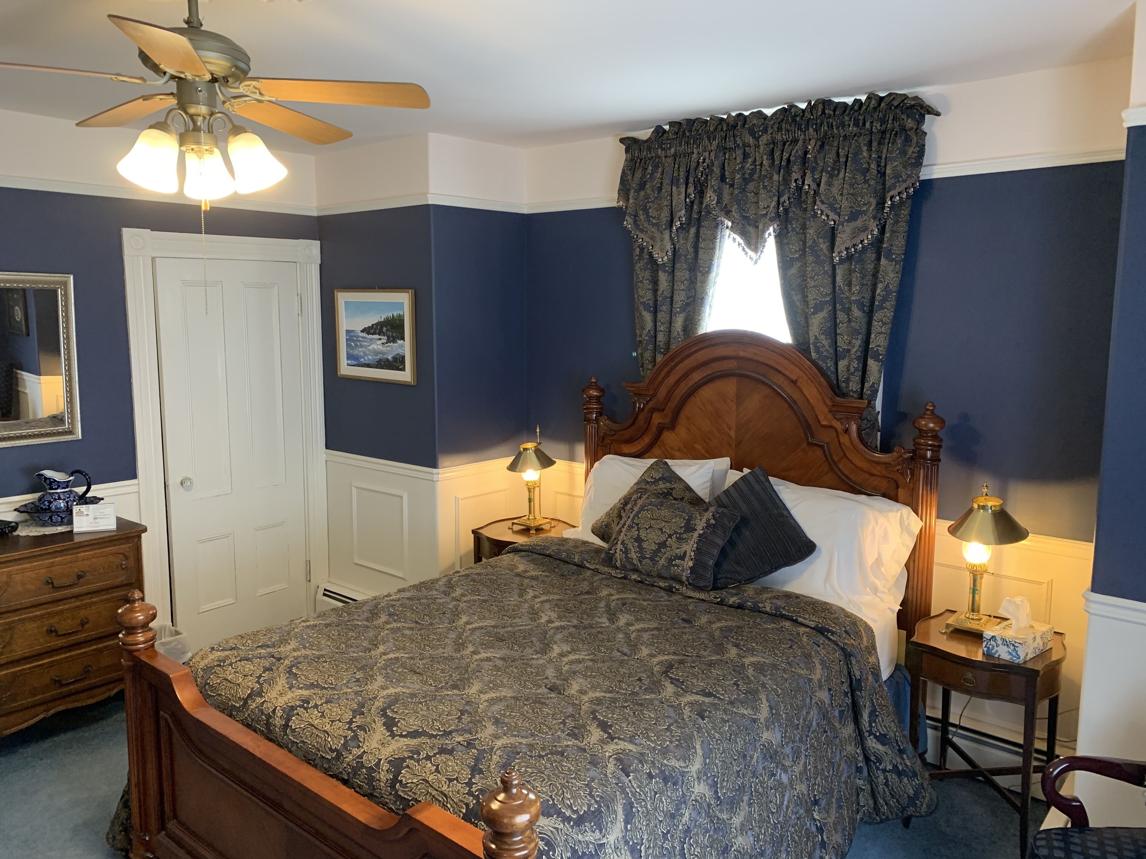Une chambre à coucher avec un lit en bois, deux tables de chevet avec des lampes, un ventilateur de plafond, une commode, une porte et une fenêtre avec des rideaux.