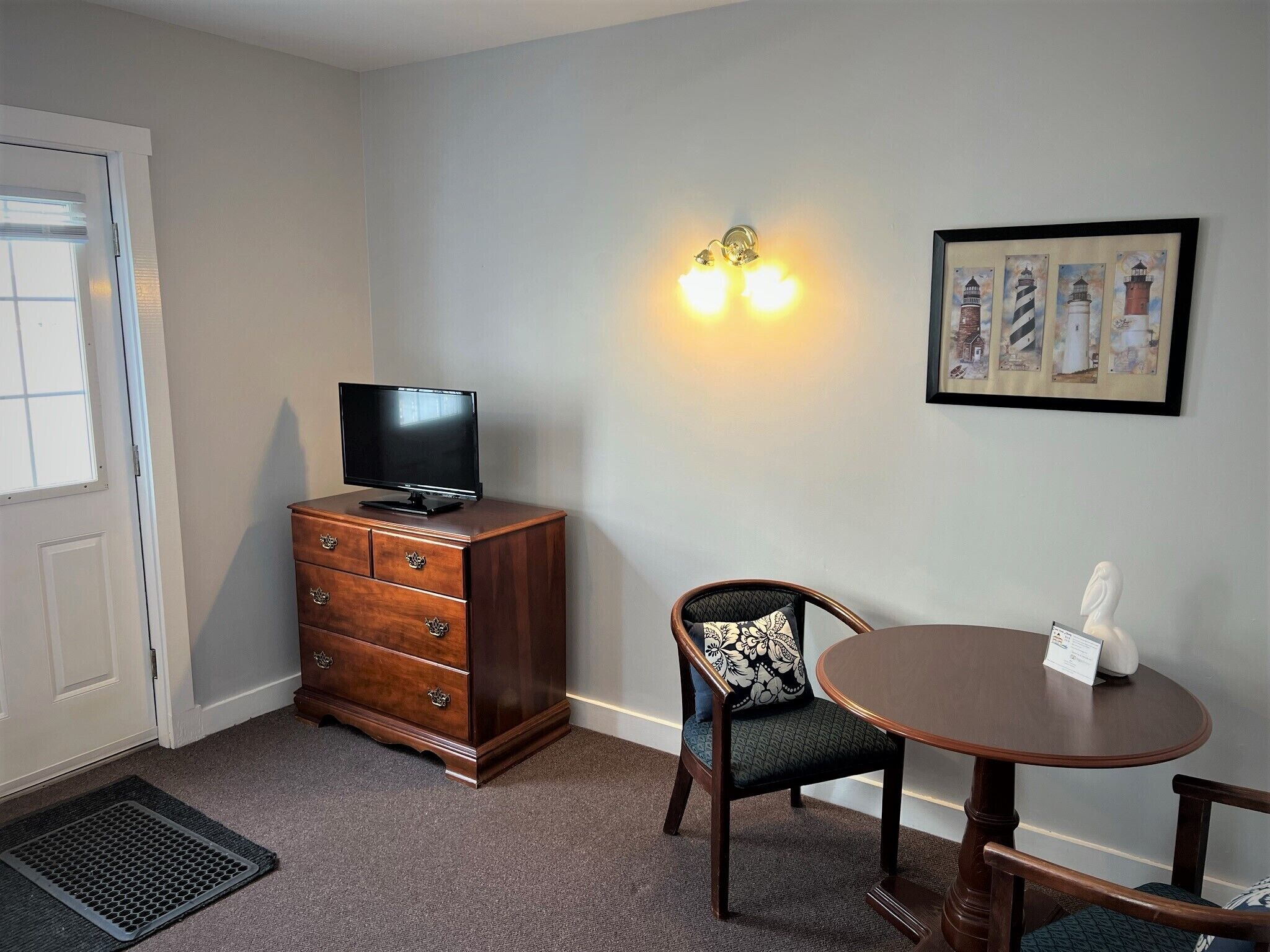 A room with a wooden dresser, a television, a chair, a table, and a wall-mounted light fixture.