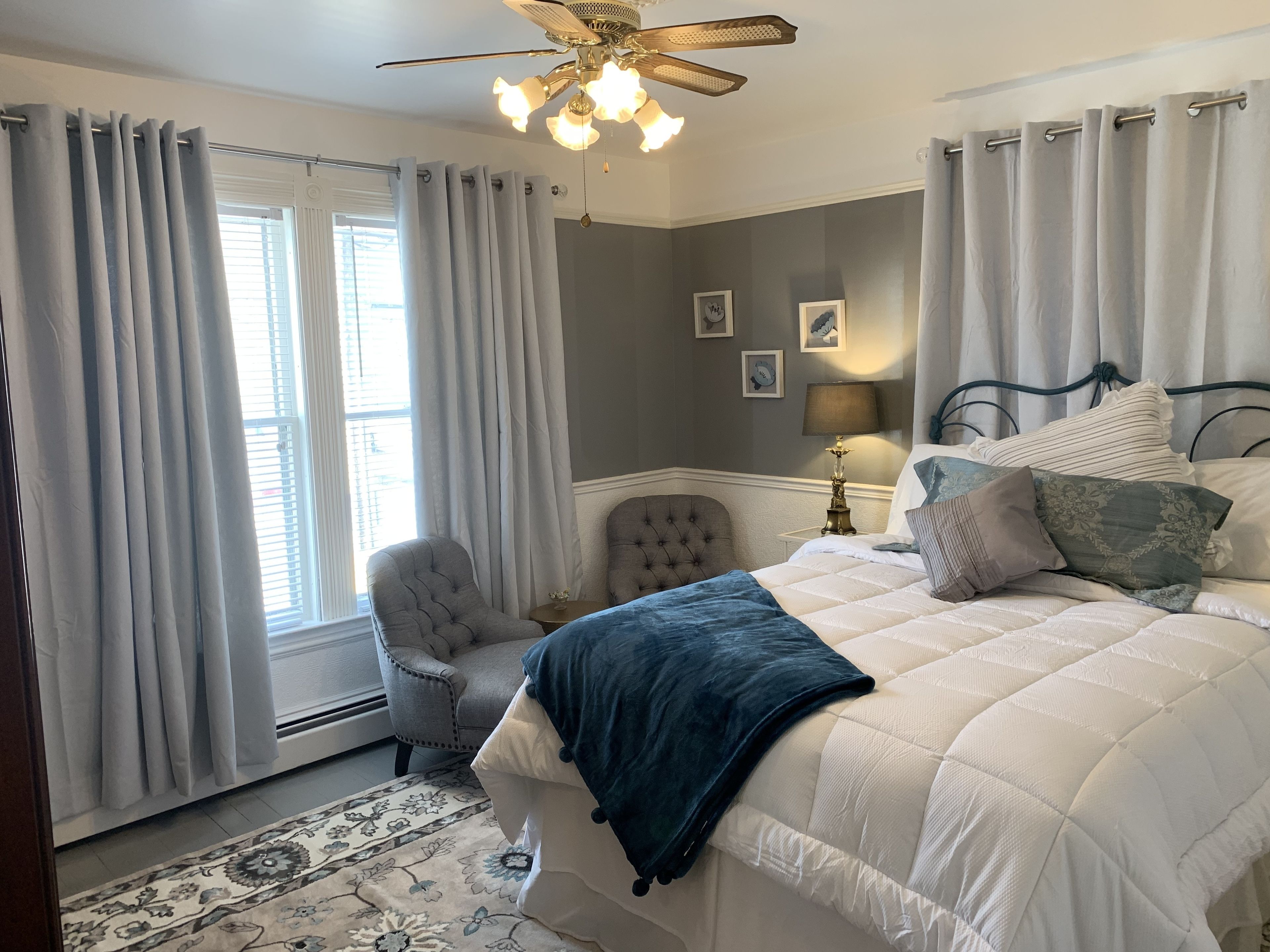 A bedroom with a bed, two chairs, a nightstand, a lamp, and a ceiling fan.