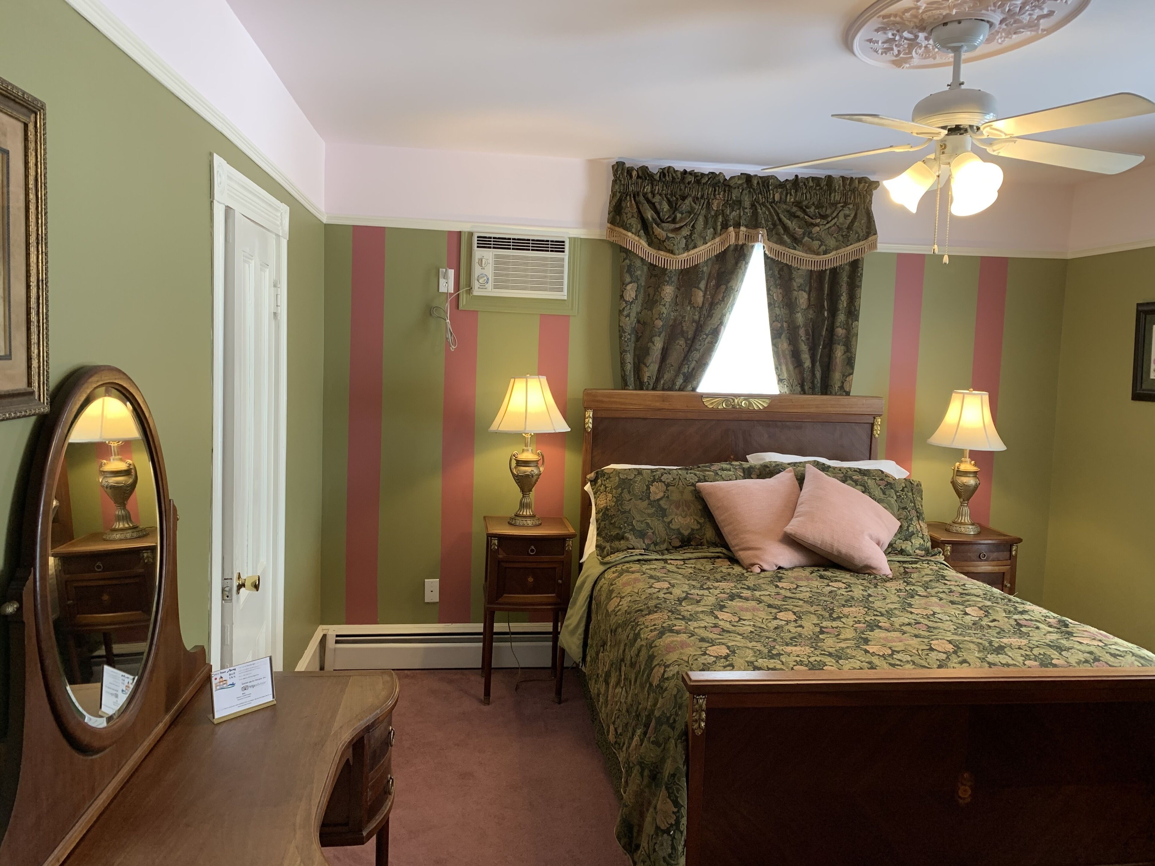 Een slaapkamer met een bed, twee nachtkastjes met lampen, een plafondventilator, een raam met gordijnen, en een wandairconditioner.