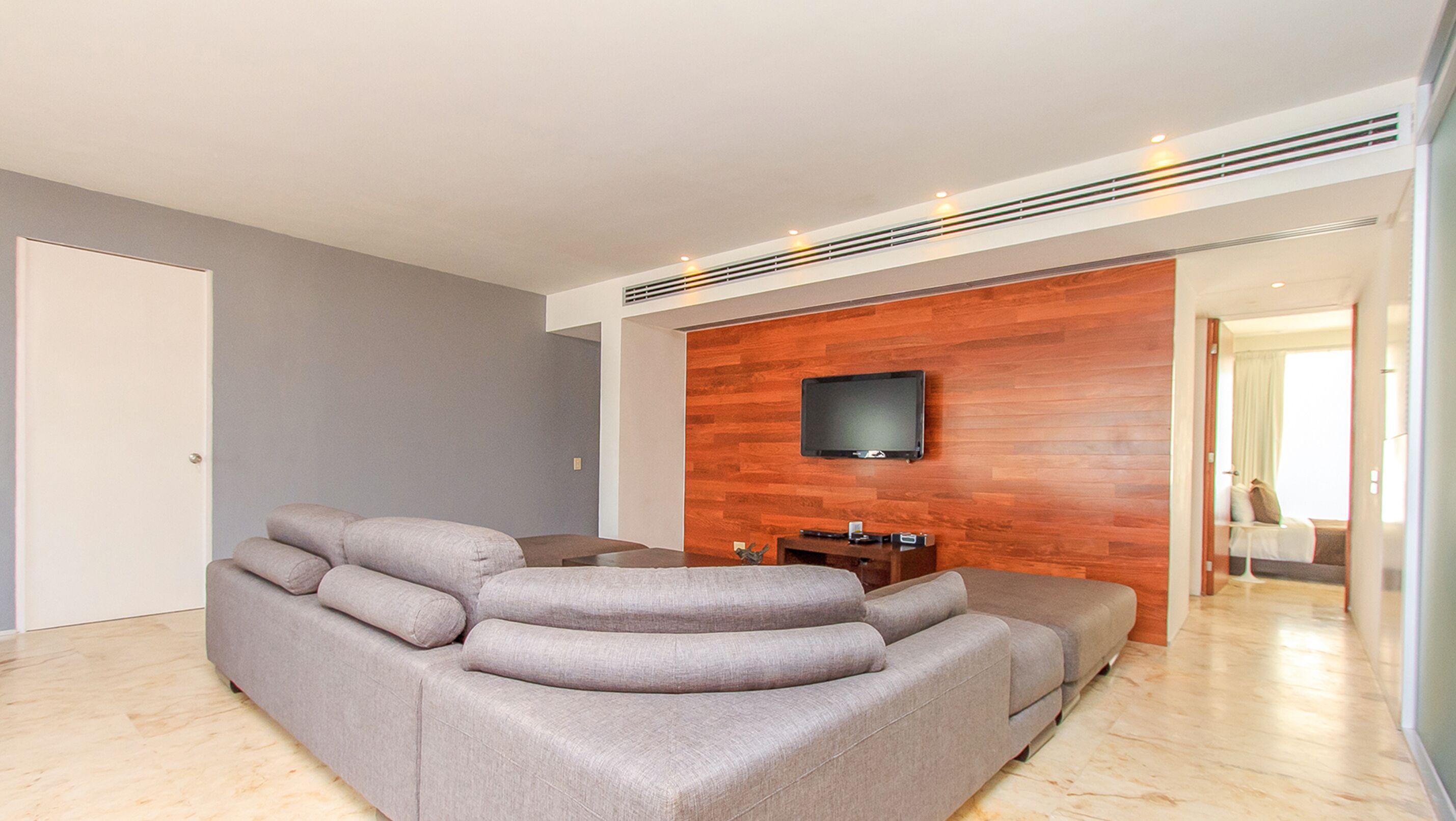 A modern living room with a grey sectional sofa, a flat-screen TV mounted on a wooden wall, and a wooden floor.