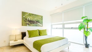 Une chambre moderne avec un grand lit, un plaid vert, un coussin vert, une table de chevet avec une lampe, un tableau abstrait accroché au mur et une plante en pot près de la fenêtre.