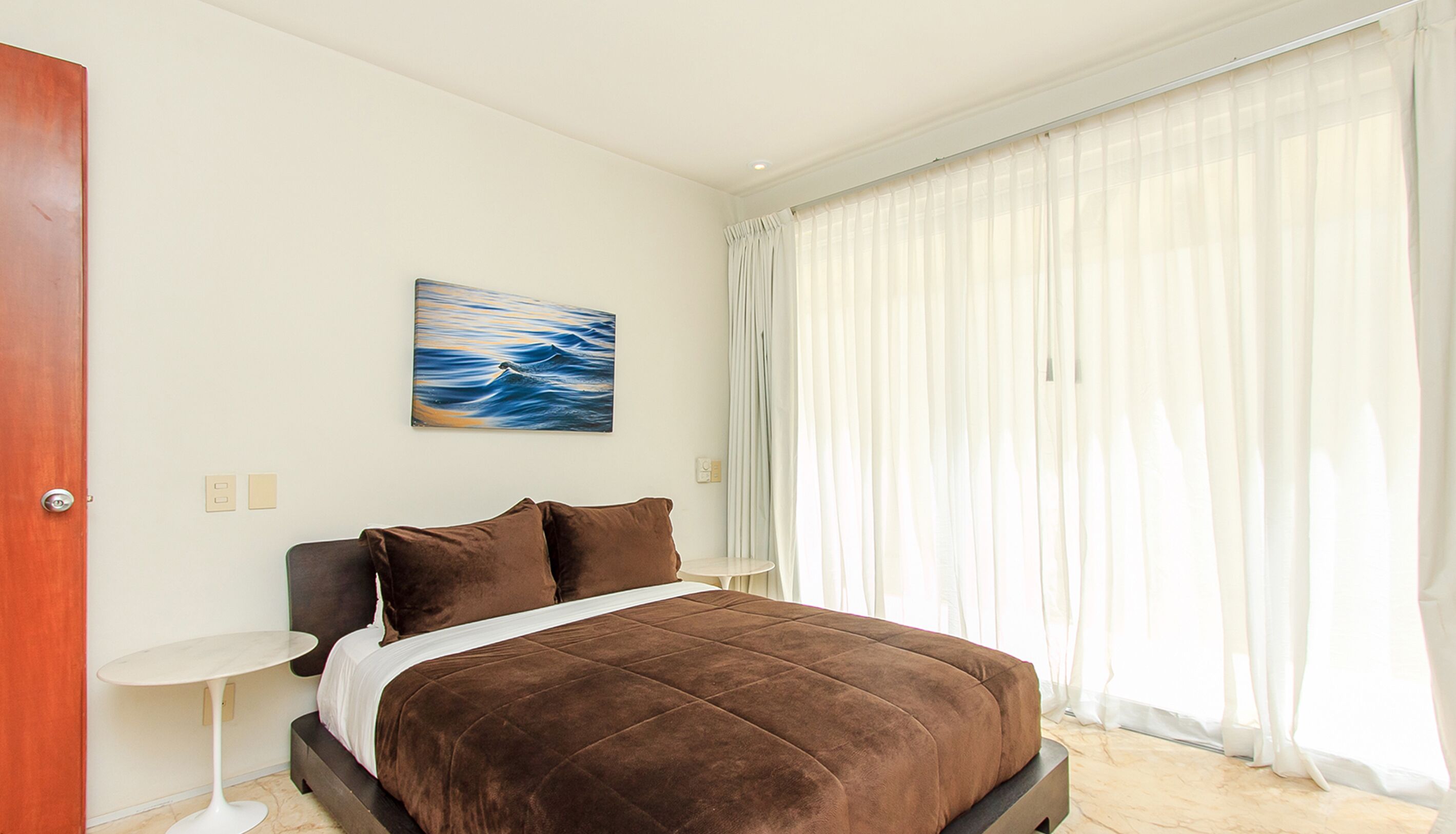 Un dormitorio con una cama, un cuadro, una ventana con cortinas y una puerta.
