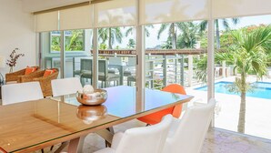 A modern dining area with a glass table, white chairs, and a view of a pool and palm trees. - MAGIA by Stella Rentals (Playa del Carmen)