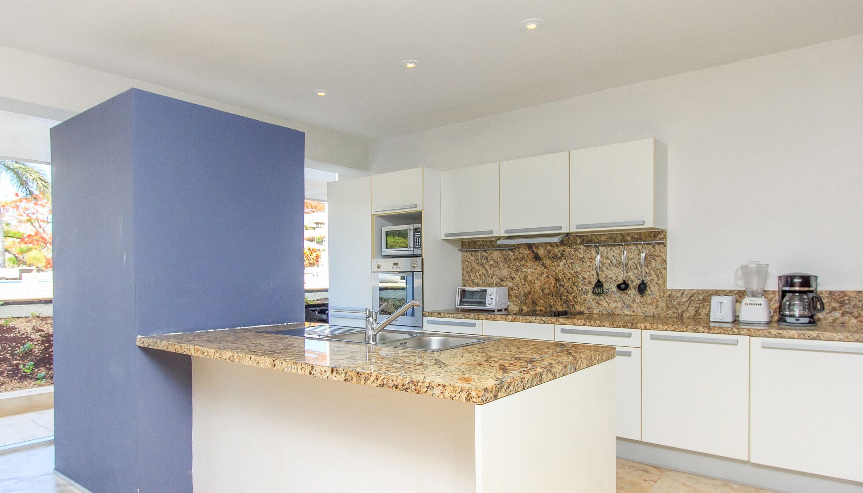 Una cocina moderna con armarios blancos, encimeras de granito y electrodomésticos de acero inoxidable, incluyendo un microondas, horno y cafetera.