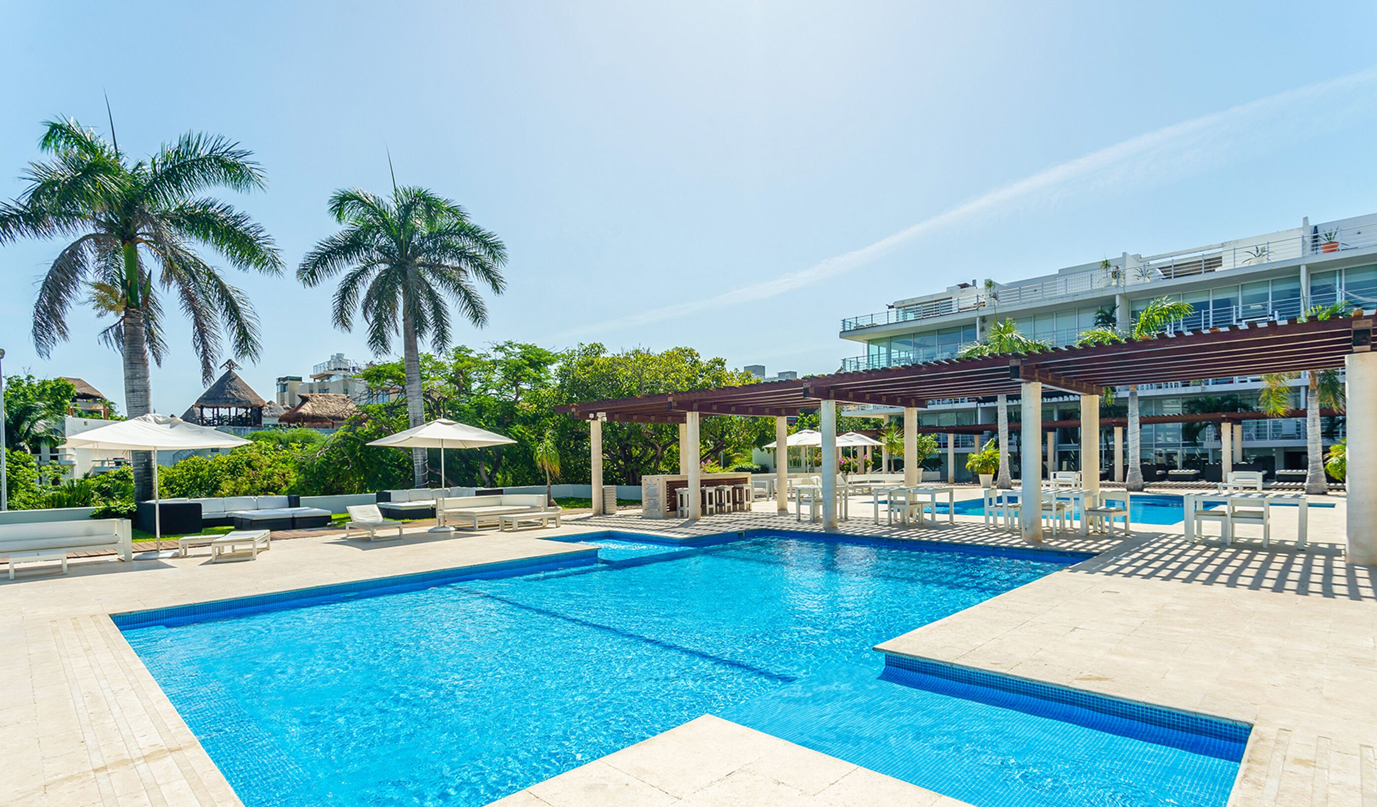 A swimming pool with lounge chairs and umbrellas, surrounded by palm trees and a building with a modern design.
