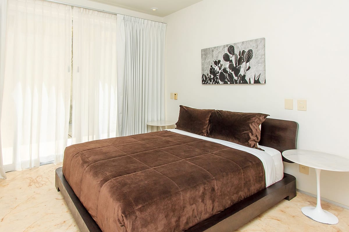Une chambre d’hôtel moderne avec un grand lit, des rideaux blancs, une petite table ronde et une œuvre d’art représentant un cactus accrochée au mur.