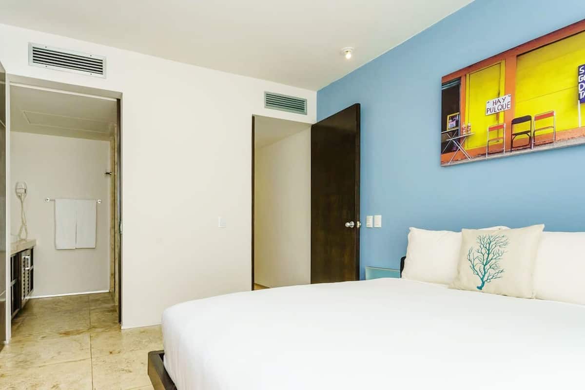 Une chambre d’hôtel avec un lit, une salle de bain où une serviette est suspendue, et une porte qui donne sur une autre pièce.