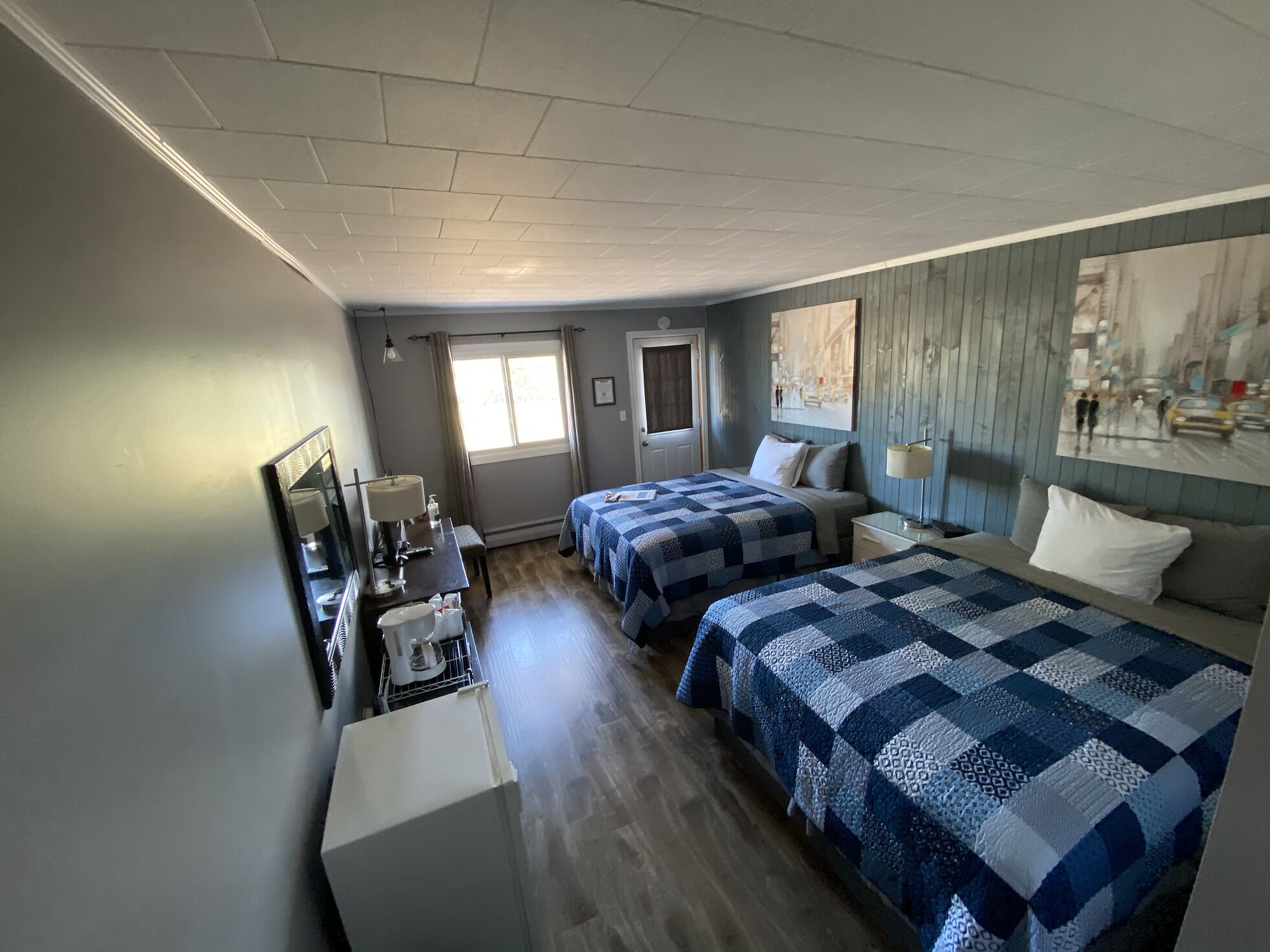 A hotel room with two beds, a desk with a lamp, a window with curtains, and a painting on the wall.