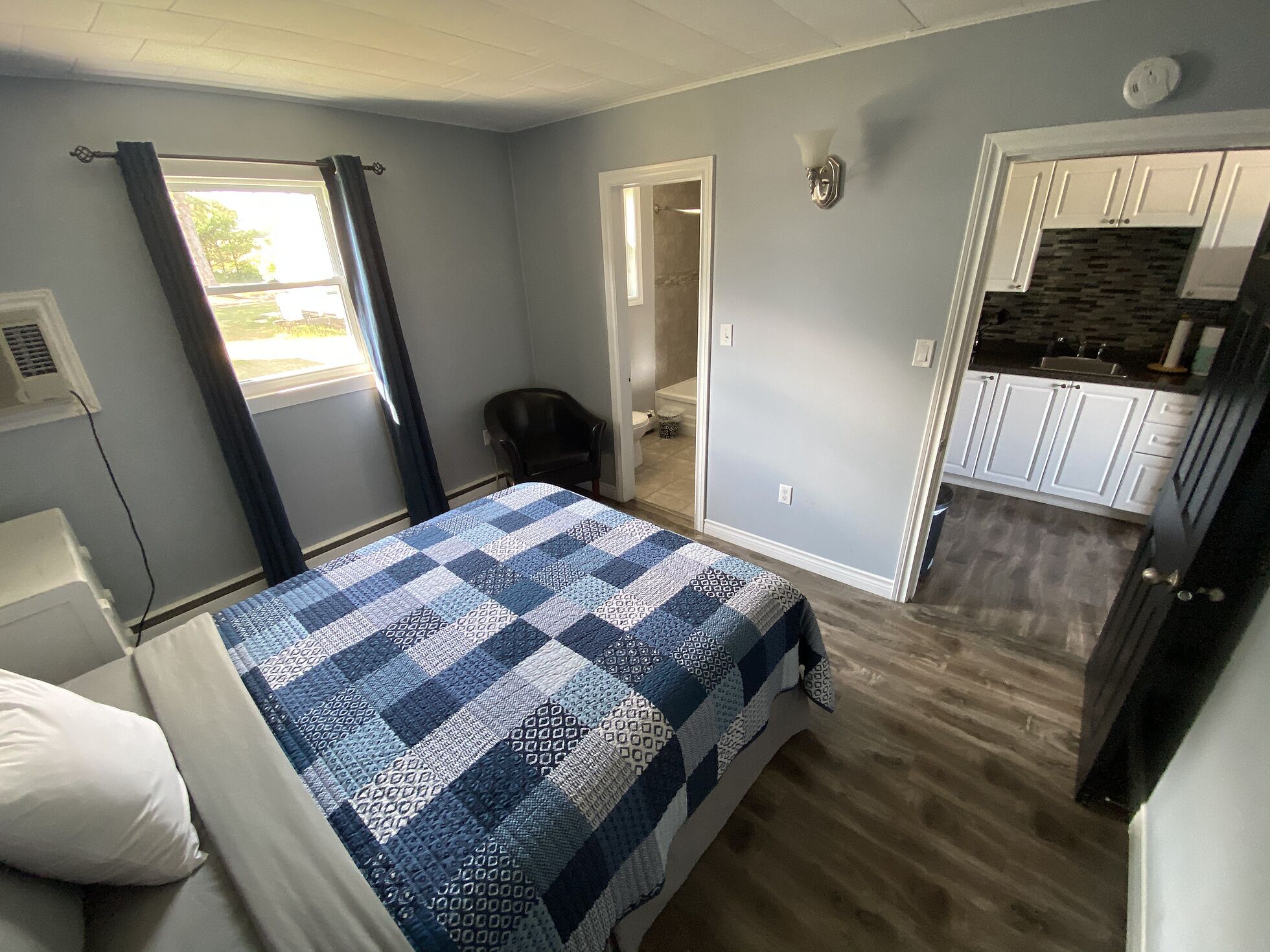 A bedroom with a bed, a chair, and a view of a bathroom and kitchen.