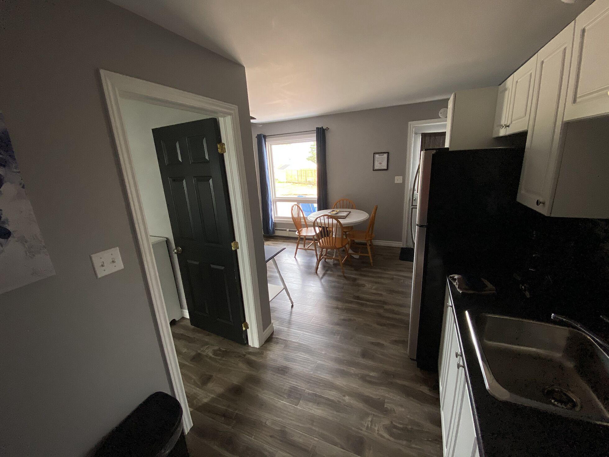Cocina con encimera negra, pileta de acero inoxidable, mesa de madera con sillas y una puerta que da a otra habitación.