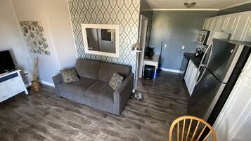 A modern living room with a gray sofa, a coffee table, and a dining area with a wooden chair. A kitchen with a refrigerator and a microwave is visible in the background.