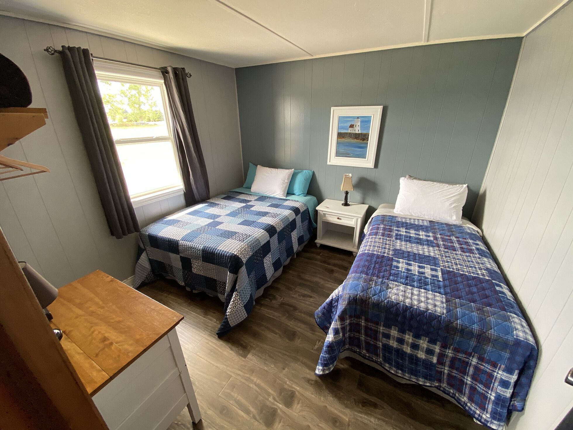 A compact bedroom with two beds, a small bedside table, a window with curtains, and a painting on the wall.