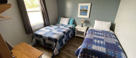A compact bedroom with two beds, a small bedside table, a window with curtains, and a painting on the wall.
