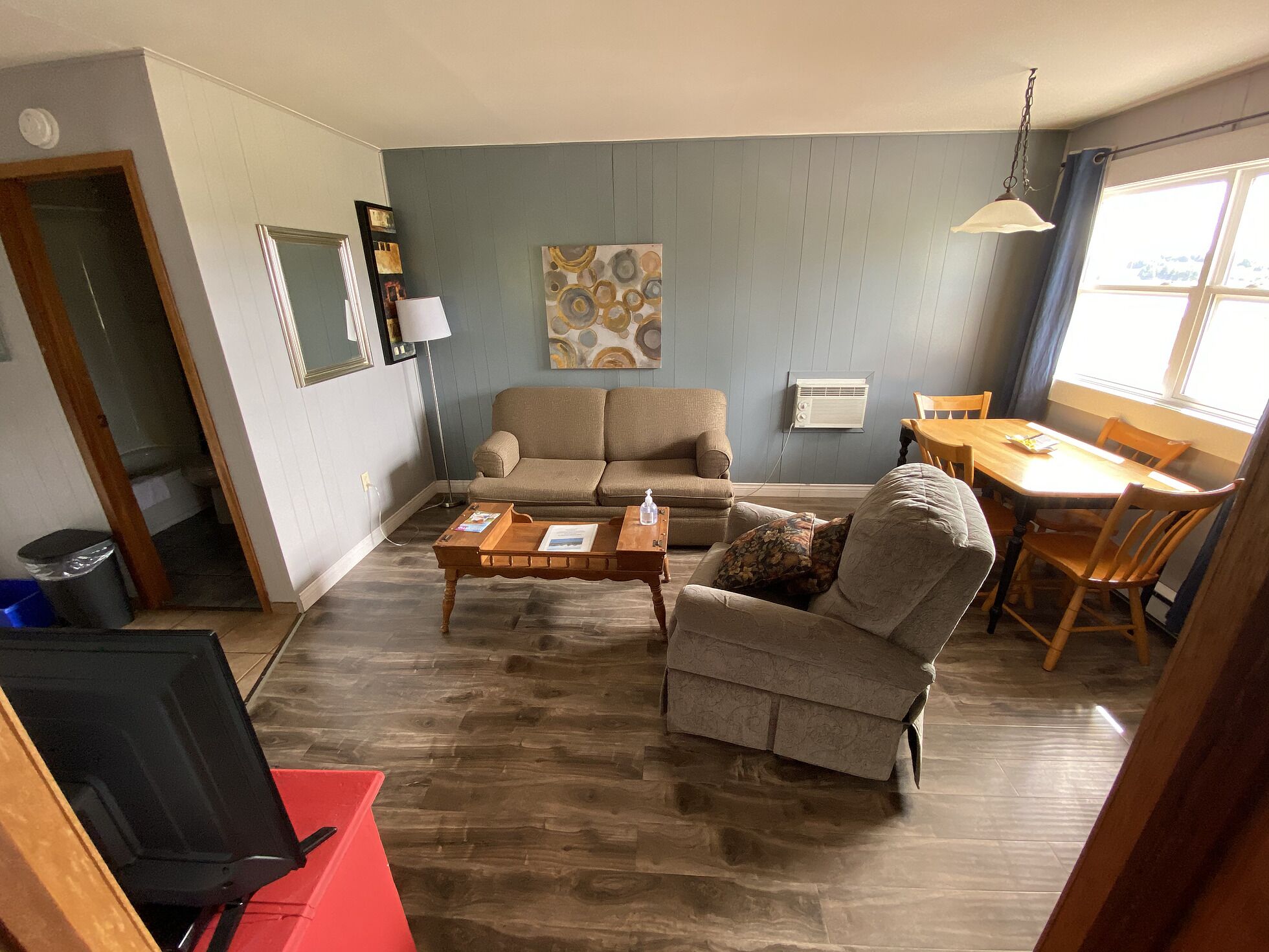 En stue med sofa, lænestol, sofabord og spisebord. Et maleri hænger på væggen, der er en lampe, og et vindue med gardiner.