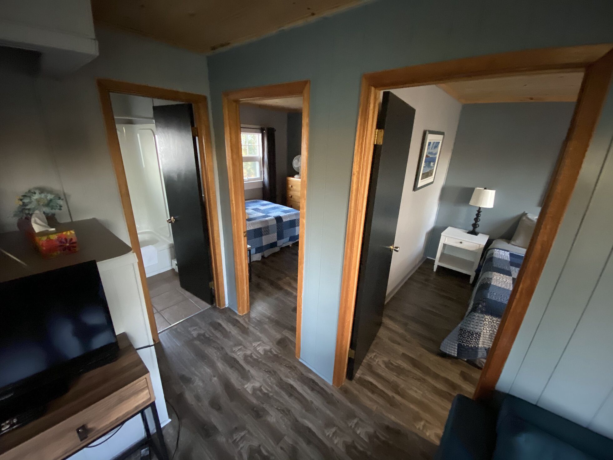 Un espace de vie compact comprenant un coin cuisine, un lit recouvert d’une couverture à carreaux, une table de chevet avec une lampe et un petit coin salon avec un coussin.