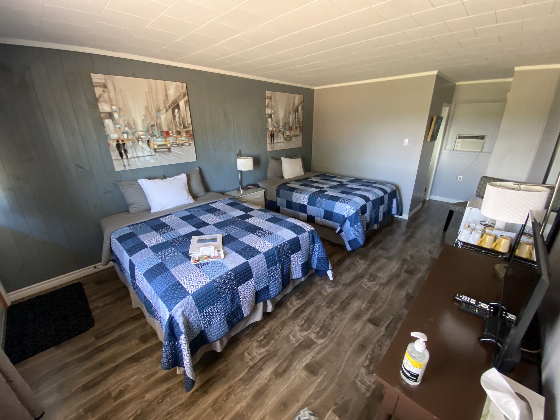 A hotel room with two beds, a desk with a lamp, a small table with toiletries, and artwork on the walls.